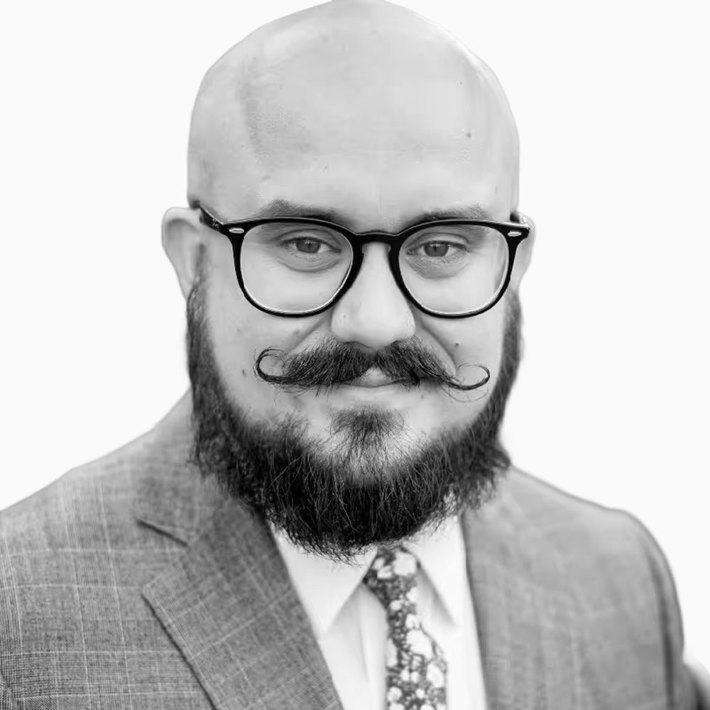 Professional black and white headshot of Jon, Account Executive at Key Outreach, wearing round glasses, a suit jacket, patterned tie, and styled mustache and beard while posing against a light gray background.