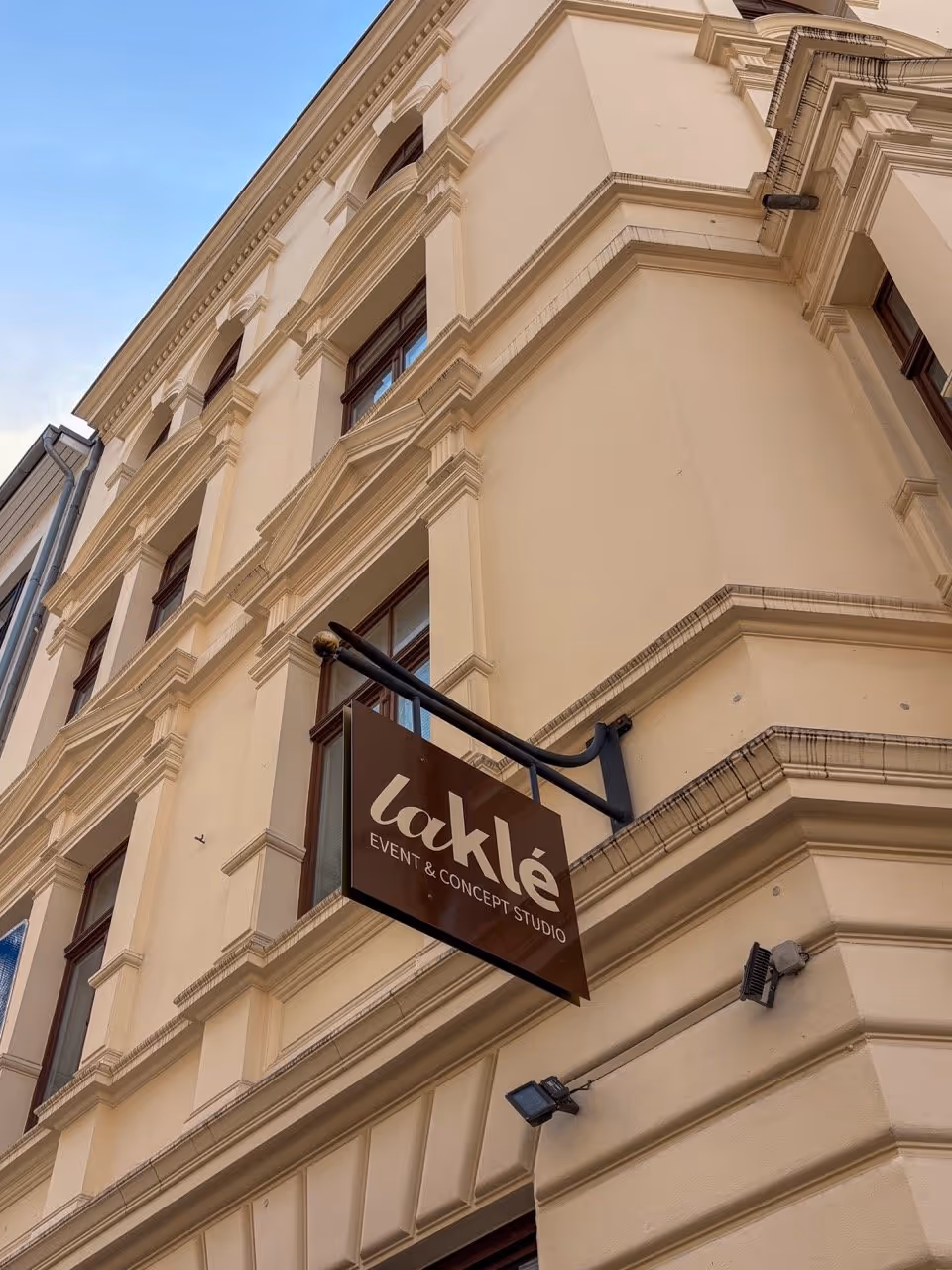 Brown sign reading 'laklé EVENT & CONCEPT STUDIO' hangs on beige decorative building corner.