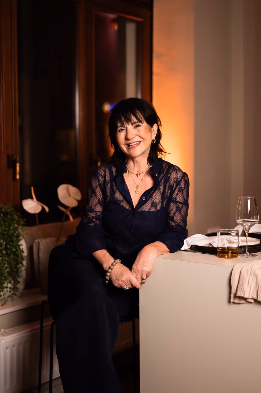 Smiling woman in a dark lace top sitting indoors near a table with glasses and a window behind her.
