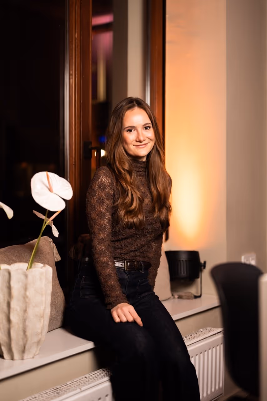 Young woman with long brown hair sits on a windowsill smiling, wearing a brown lace top and black pants.