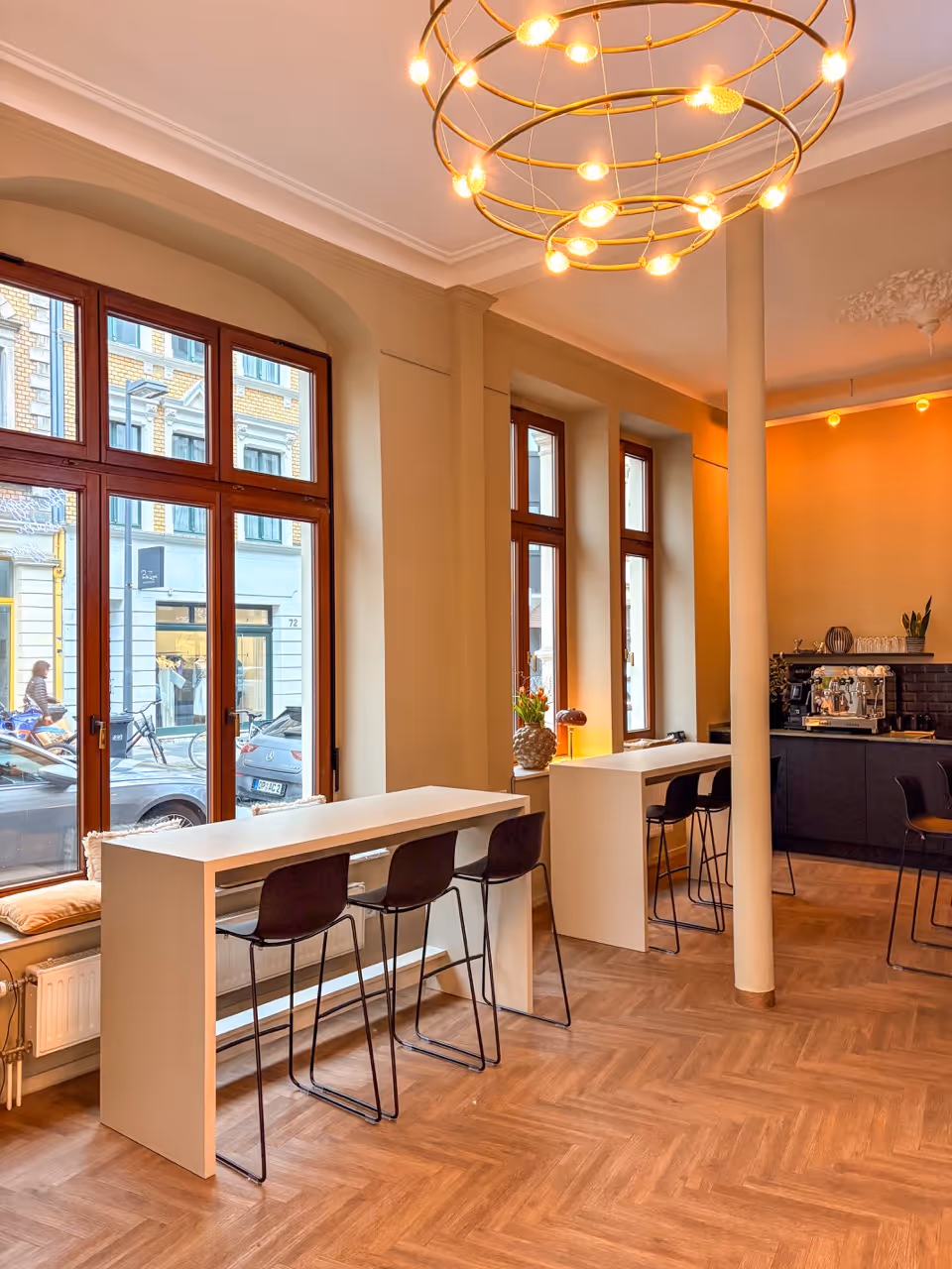 Modern cafe interior with wooden floors, tall white tables, black chairs, and large windows.
