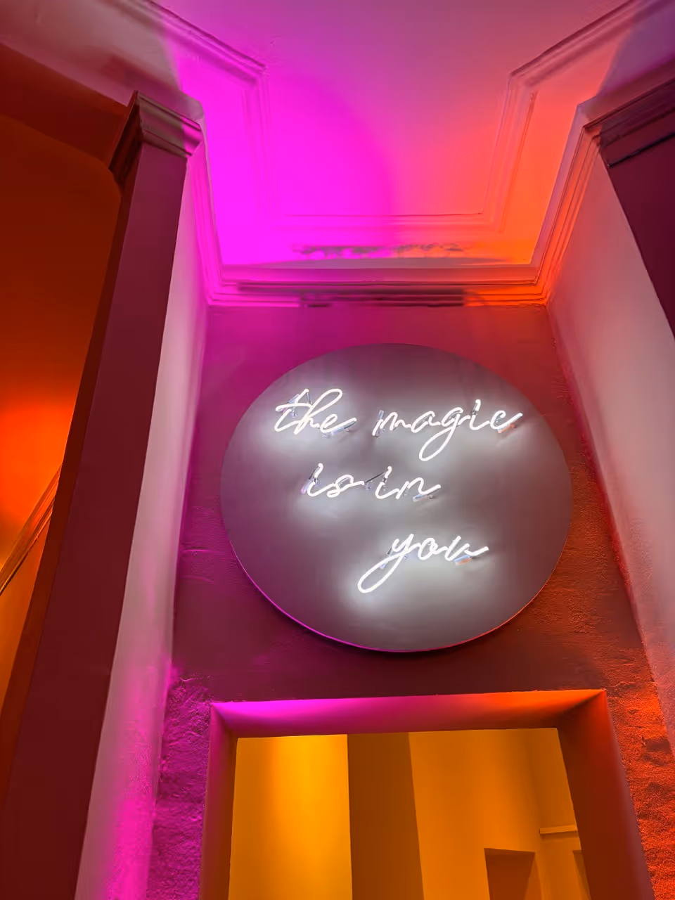 Circular neon sign reading 'the magic is in you' with pink and orange light on surrounding walls.