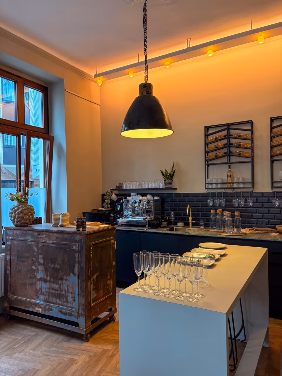Modern kitchen with a large hanging lamp, a rustic wooden cart, and glasses on a white island counter.