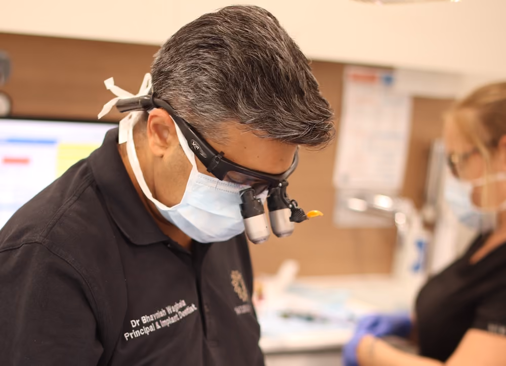 Man wearing magnifying glasses and face mask, focusing on a task, with a blurred person in the background.