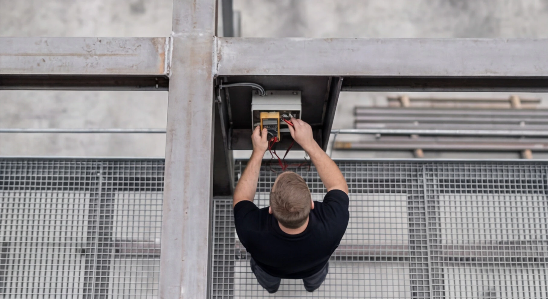 Technician in a black shirt testing electrical wiring with a multimeter on metal grating floor.