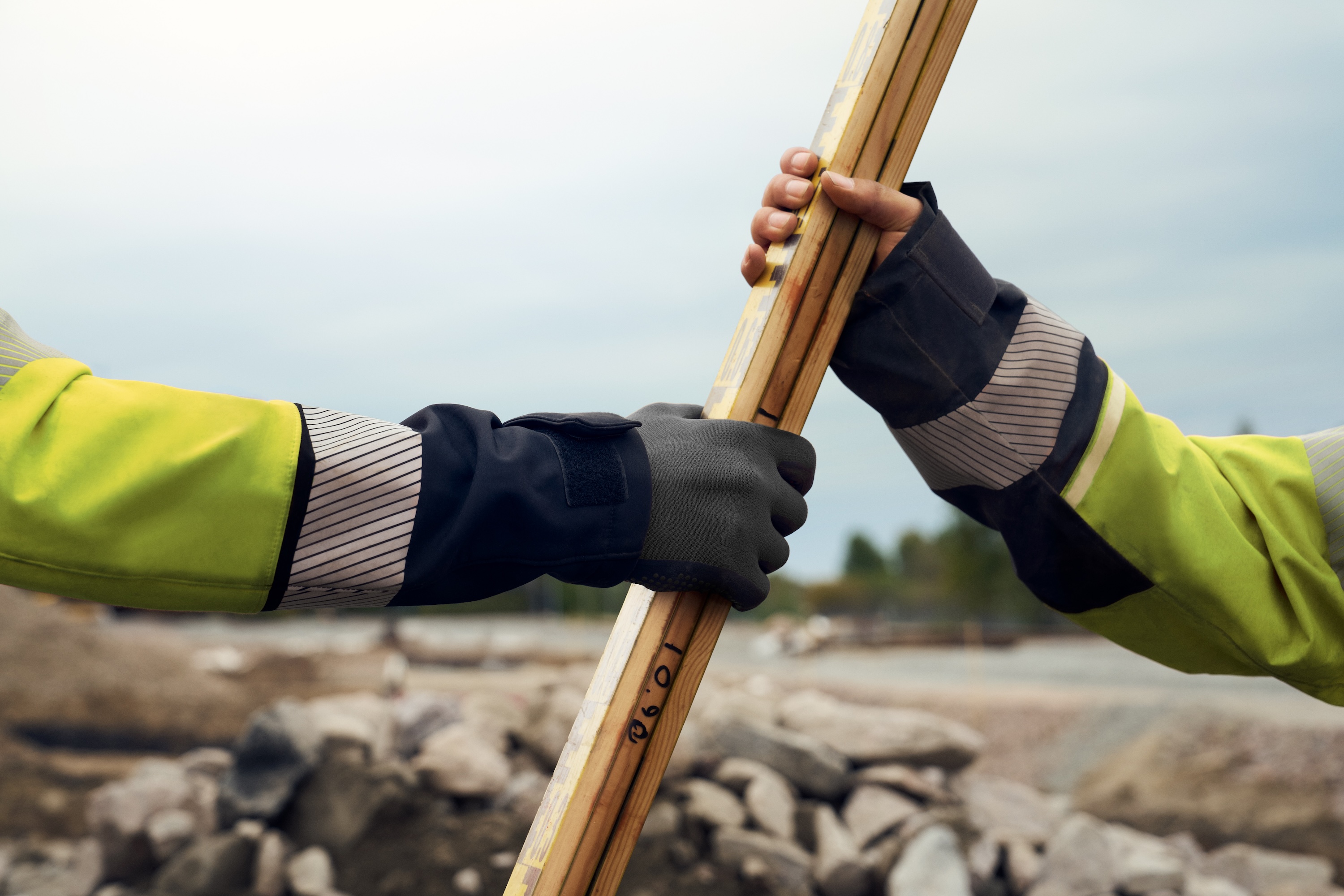 Two people in work clothes hold a measuring tool over rocky ground.