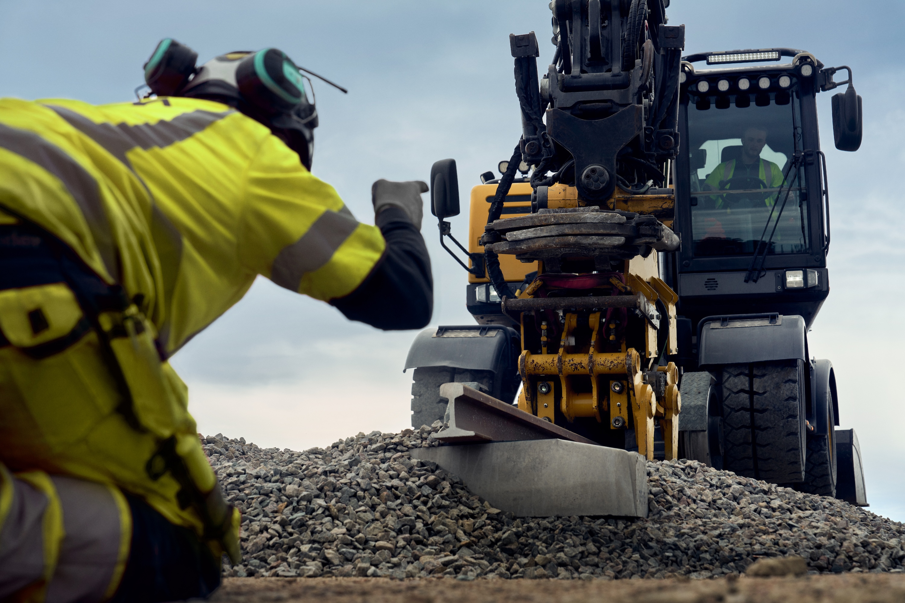 Construction worker in yellow warning jacket guides excavator over gravel at construction site.