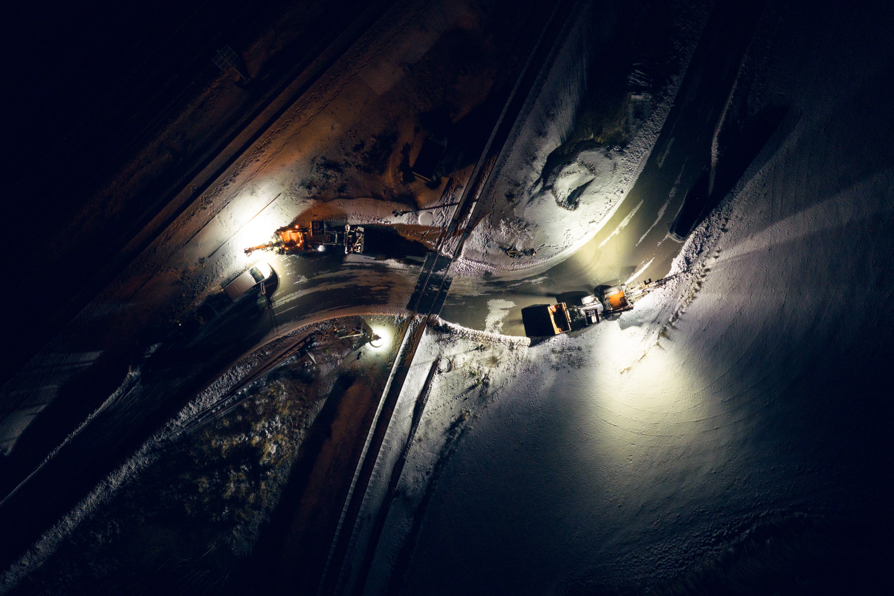 Night view of snow covered road with two snow plows and vehicles working under street lights.