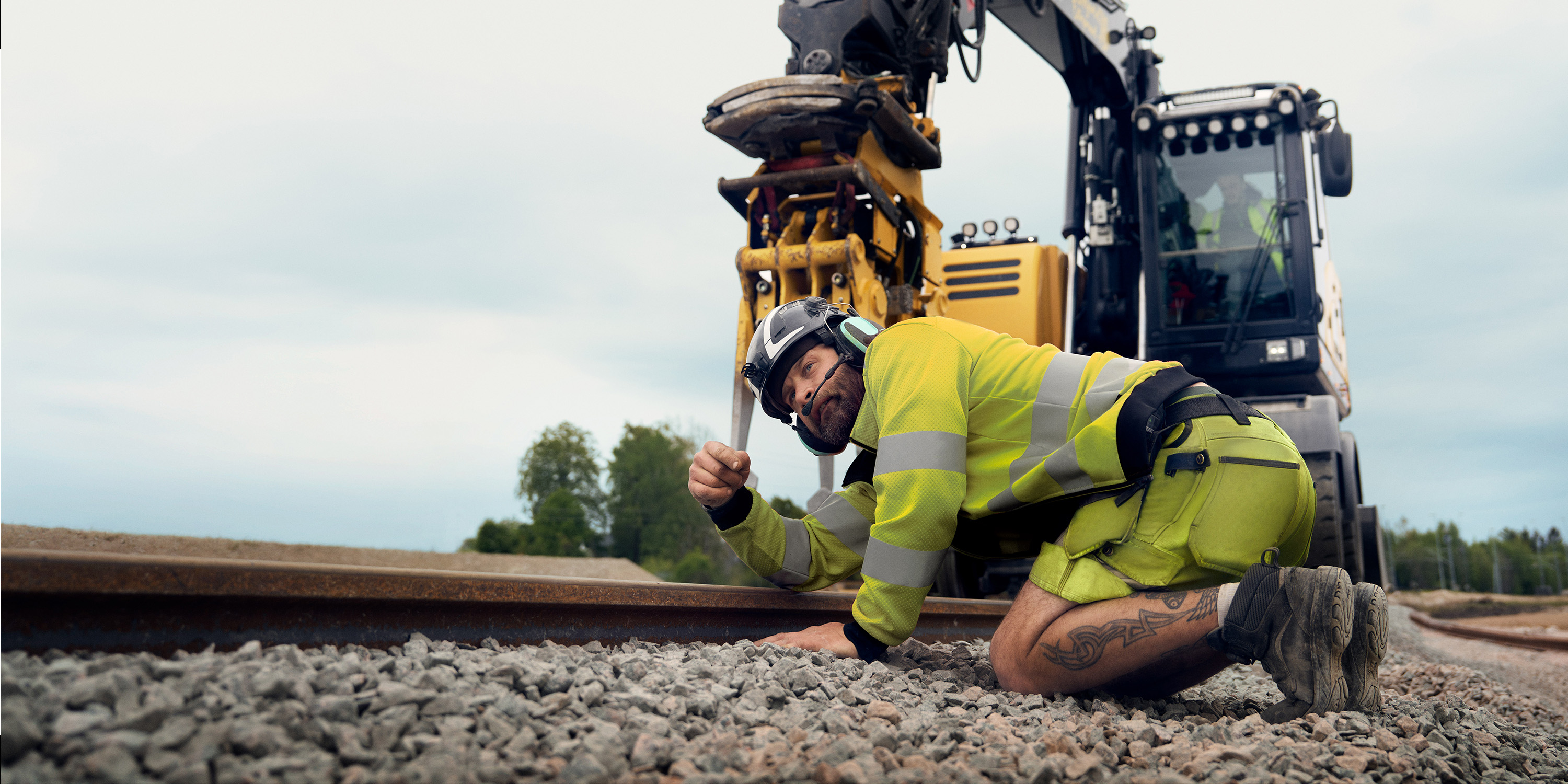 A track technician in yellow warning attire inspects railway tracks in front of a digger.