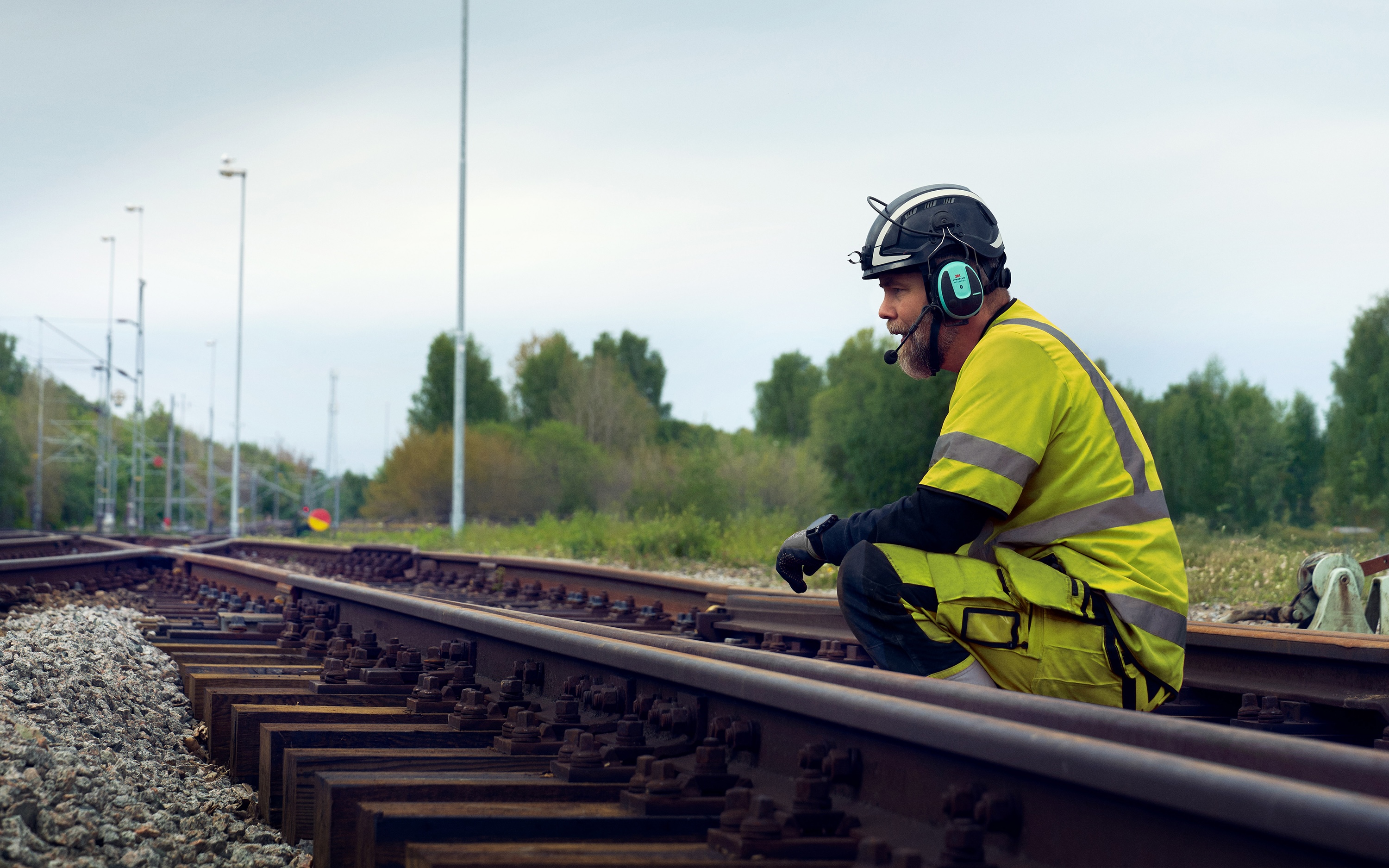 A man in warning clothes and helmet with hearing protection sits on the railway track inspecting it.