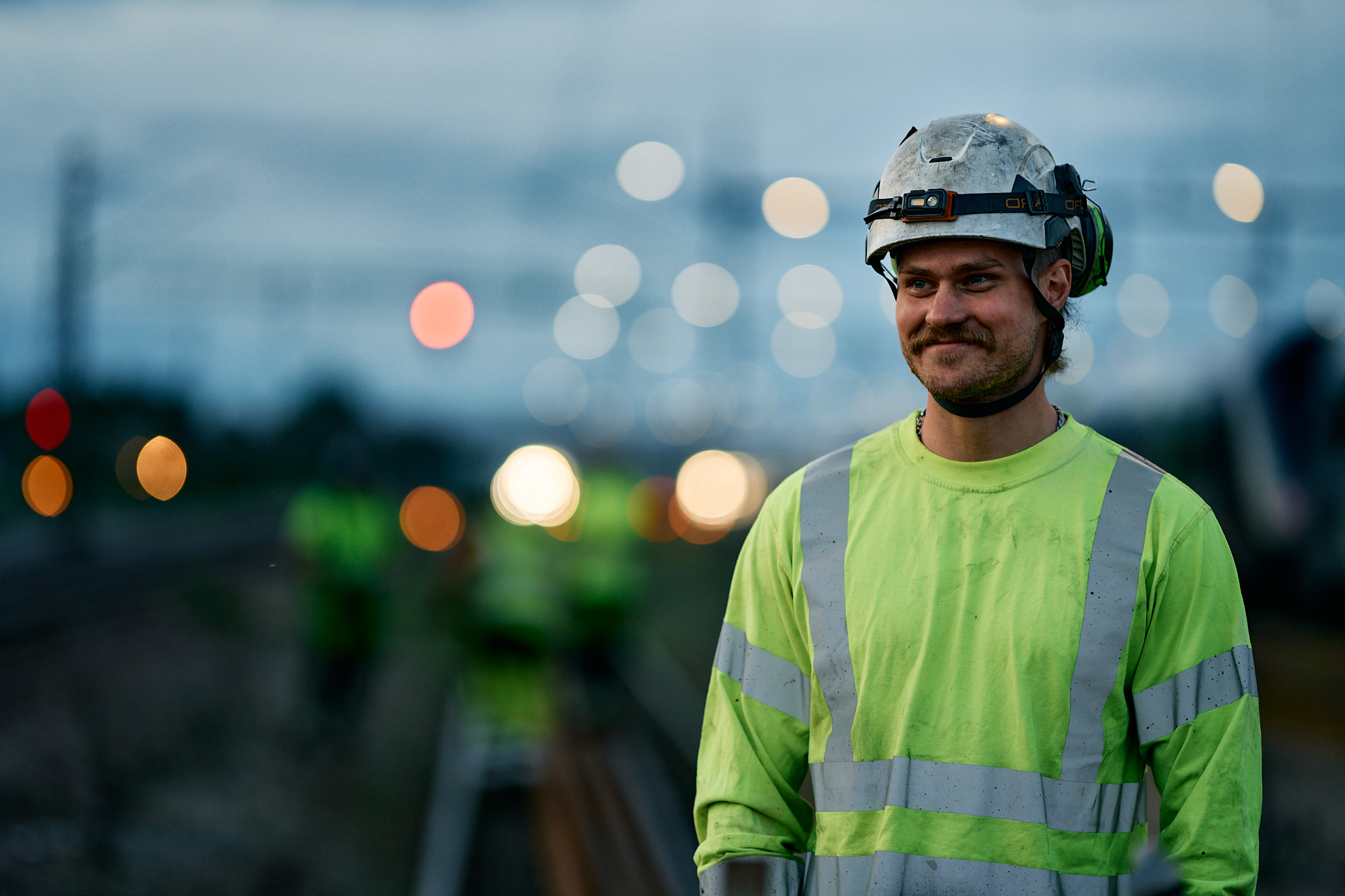 Track technician in reflective sweatshirt and helmet smiles while standing by railroad tracks at dusk.