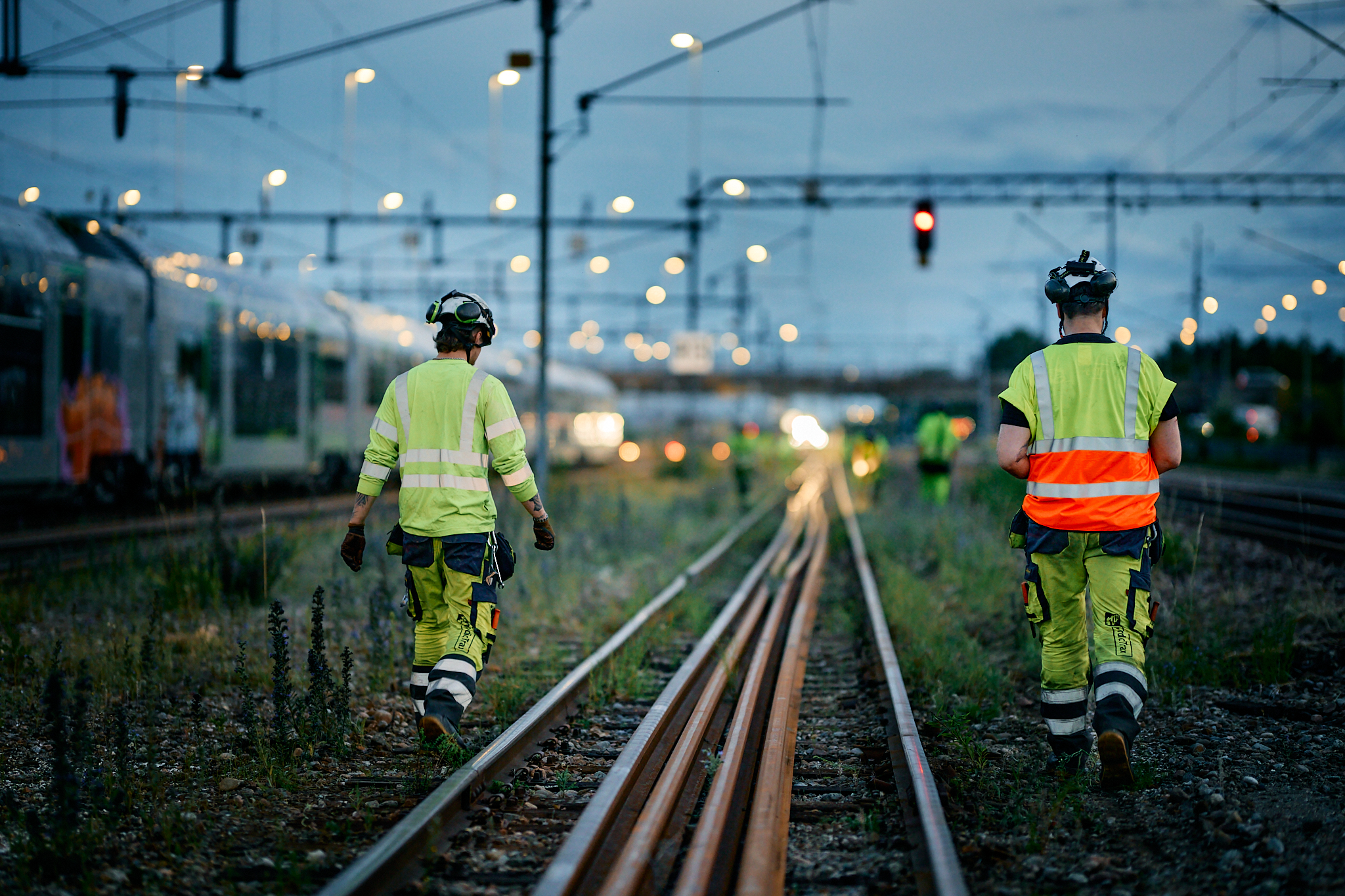 Two track technicians in reflective clothing inspect railway tracks at dusk with trains and lights in the background.