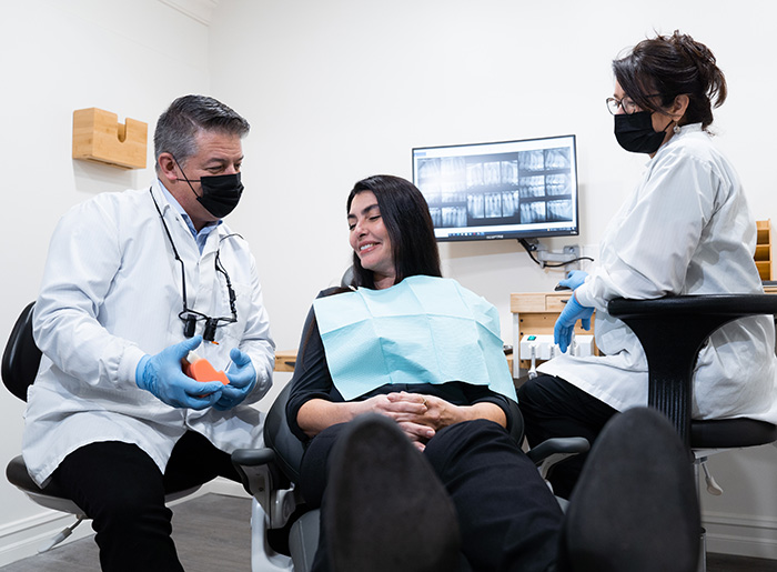 Dentist explaining dental model to a female patient in a dental chair with an assistant nearby and dental X-rays displayed on a screen behind.