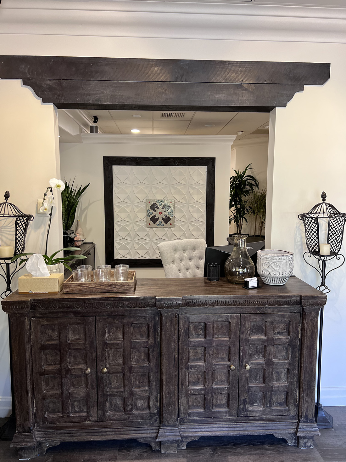 Rustic wooden reception desk with decorative items and a tufted chair behind it in a warmly lit room with wall art and plants.