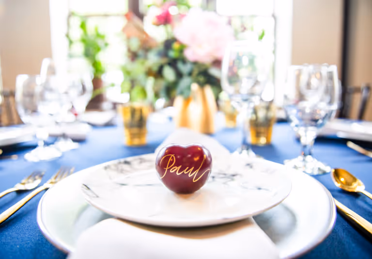 Table setting with gold cutlery, a name card reading Paul on a heart-shaped object, and blurred flowers in background.