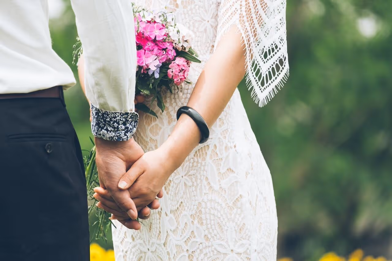 Close-up of couple holding hands, bride in lace dress holding pink flower bouquet outdoors.