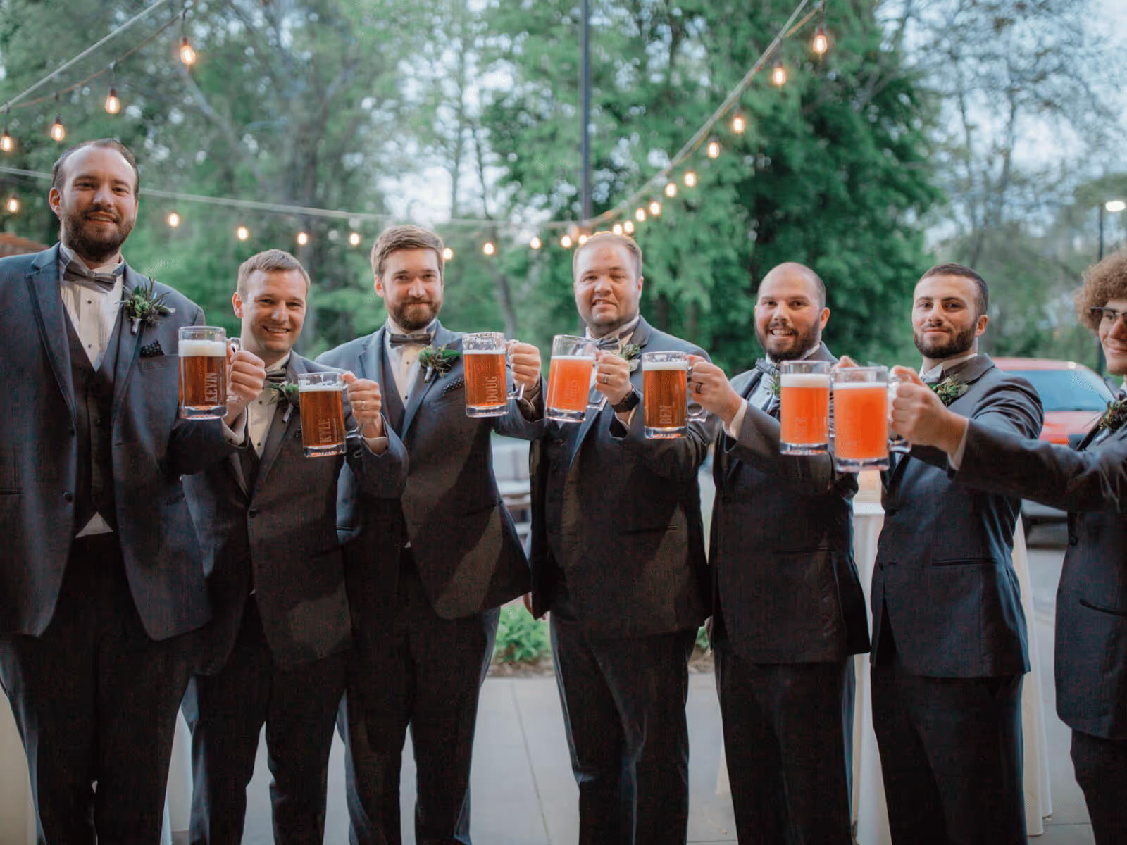 Group of men in tuxedos holding up beer mugs outdoors with string lights and trees in background.