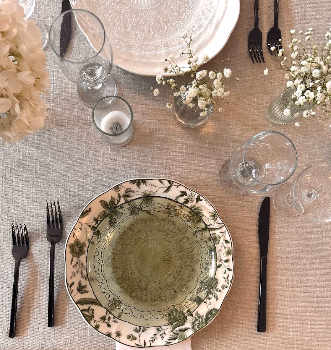 Elegant table setting with floral plates, black cutlery, glassware, candles, and small white flowers on linen.