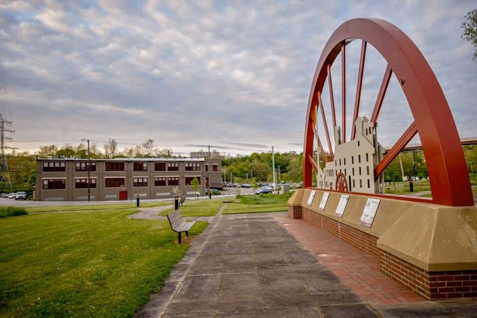 Large red wheel sculpture with plaques in a park area near a gray building and cloudy sky.