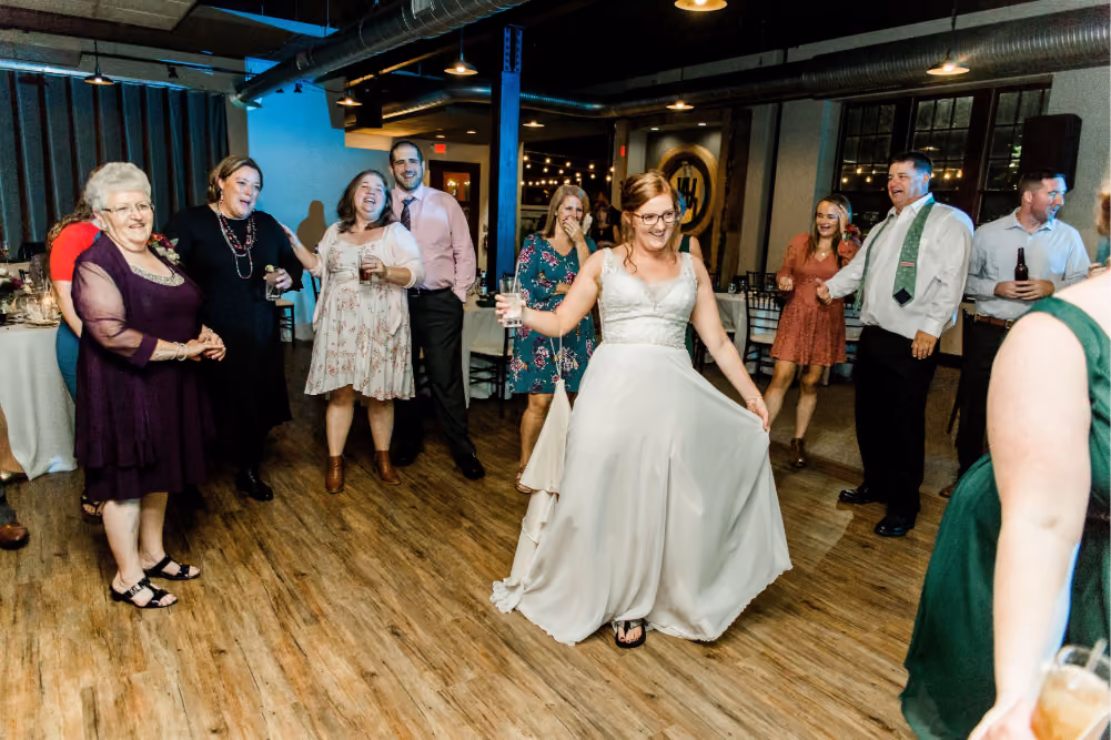Bride in white dress dancing with guests smiling and holding drinks on a wooden floor indoors.