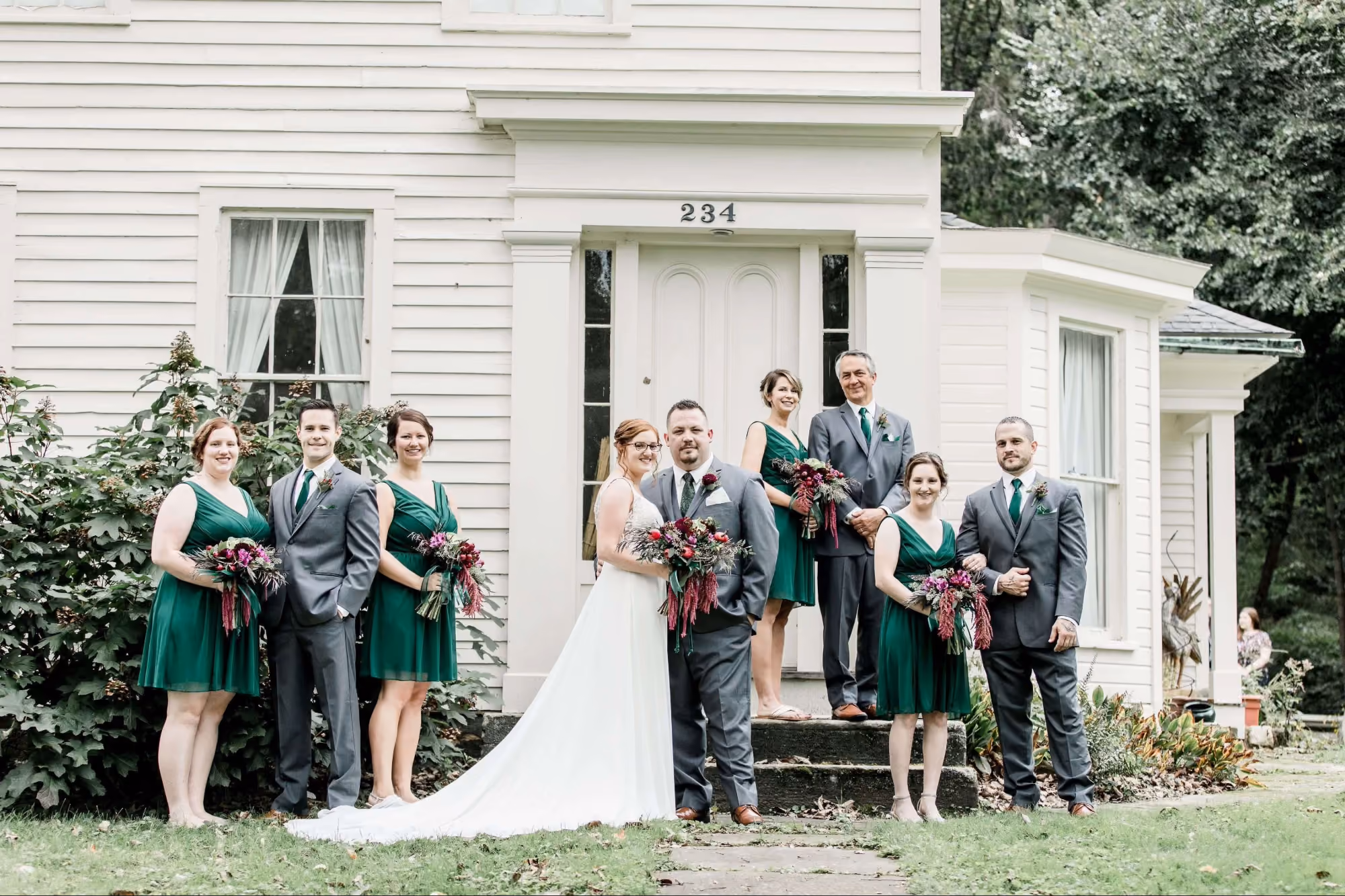 Wedding party with bride in white gown and groom in gray suit posing outside a white house.