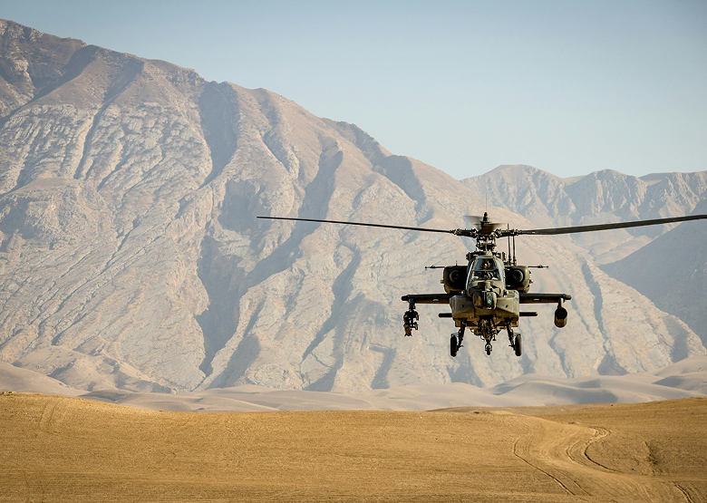 Military helicopter hovering over dry desert land with rocky mountains in the background.