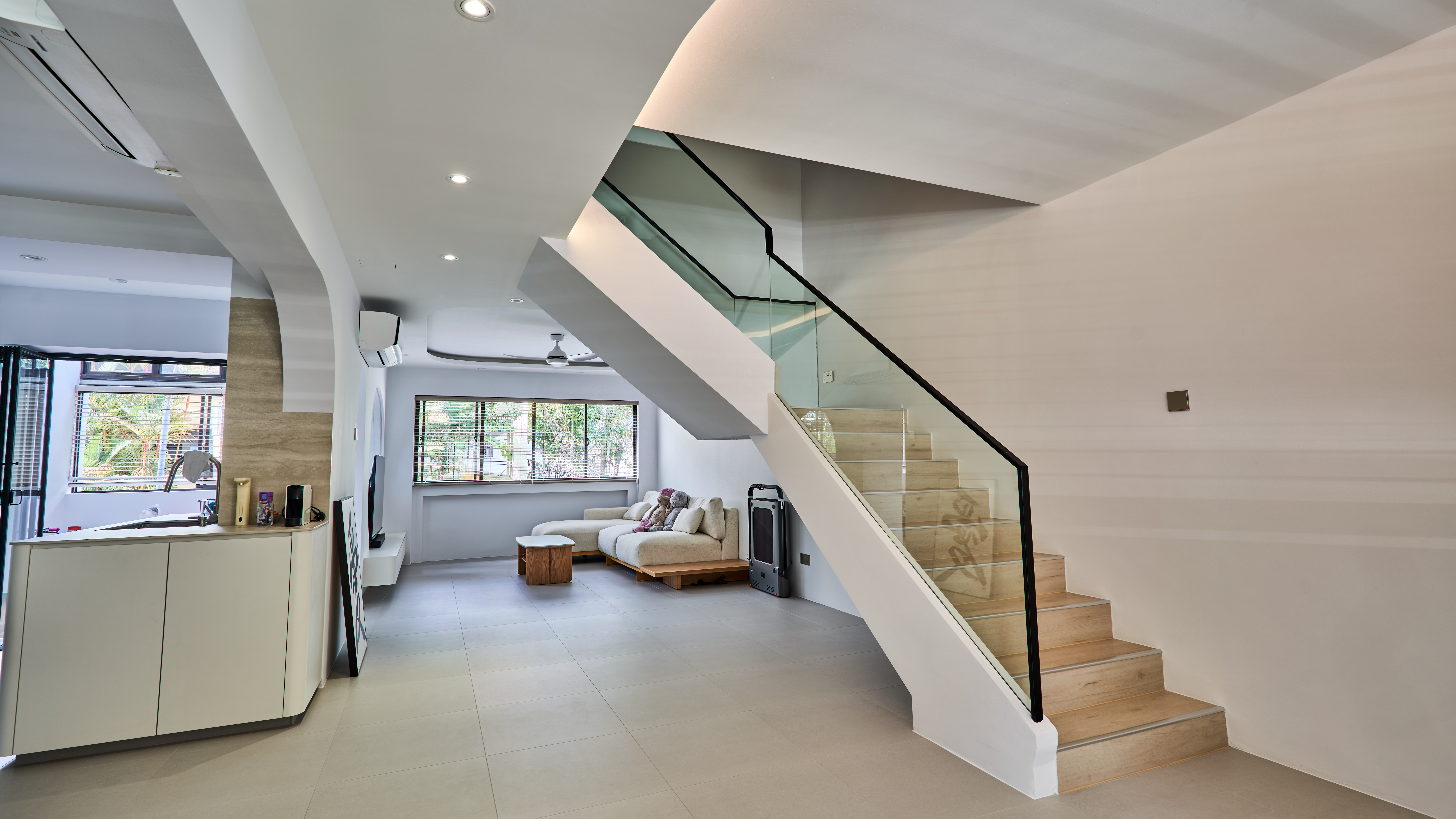 Open-plan living room with a sofa and TV console beneath a curved white staircase with glass and timber steps.