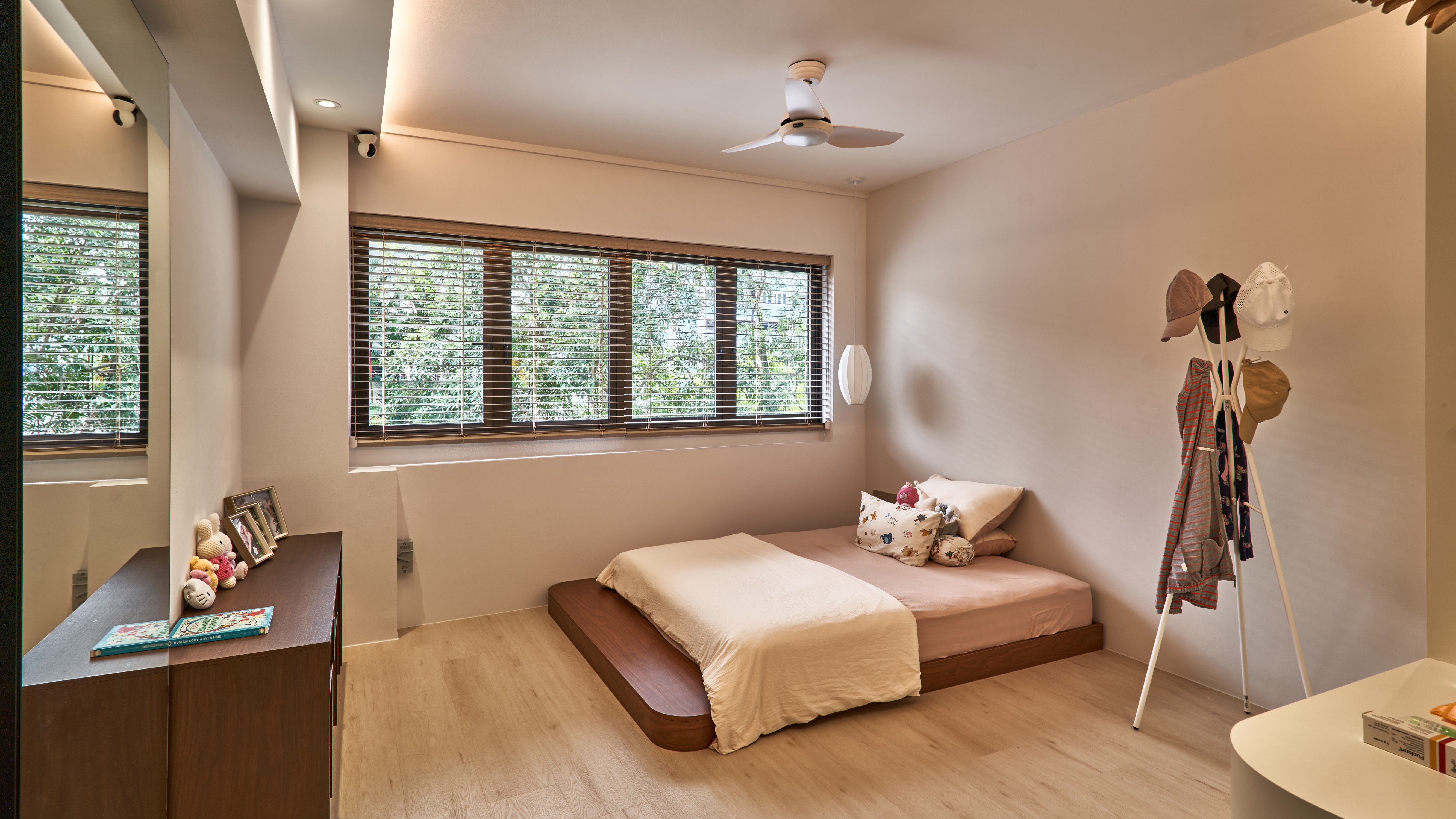 Minimalist bedroom with a low wooden platform bed, cream linens, venetian blinds, timber flooring and a wooden coat rack, with greenery visible through the windows.