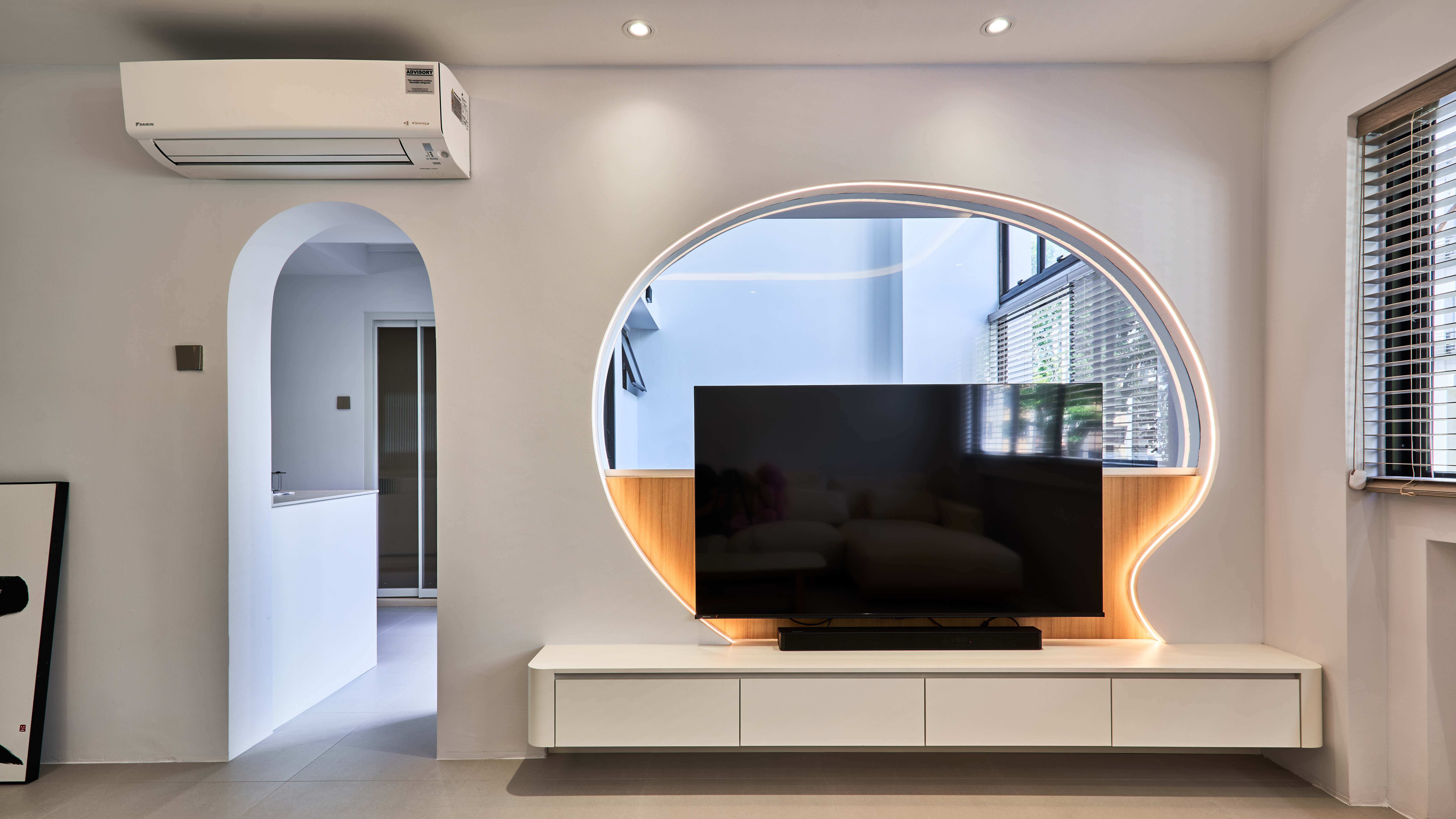 Living room with a rounded, backlit oval TV feature wall, floating white cabinet, and an arched doorway leading to the kitchen.