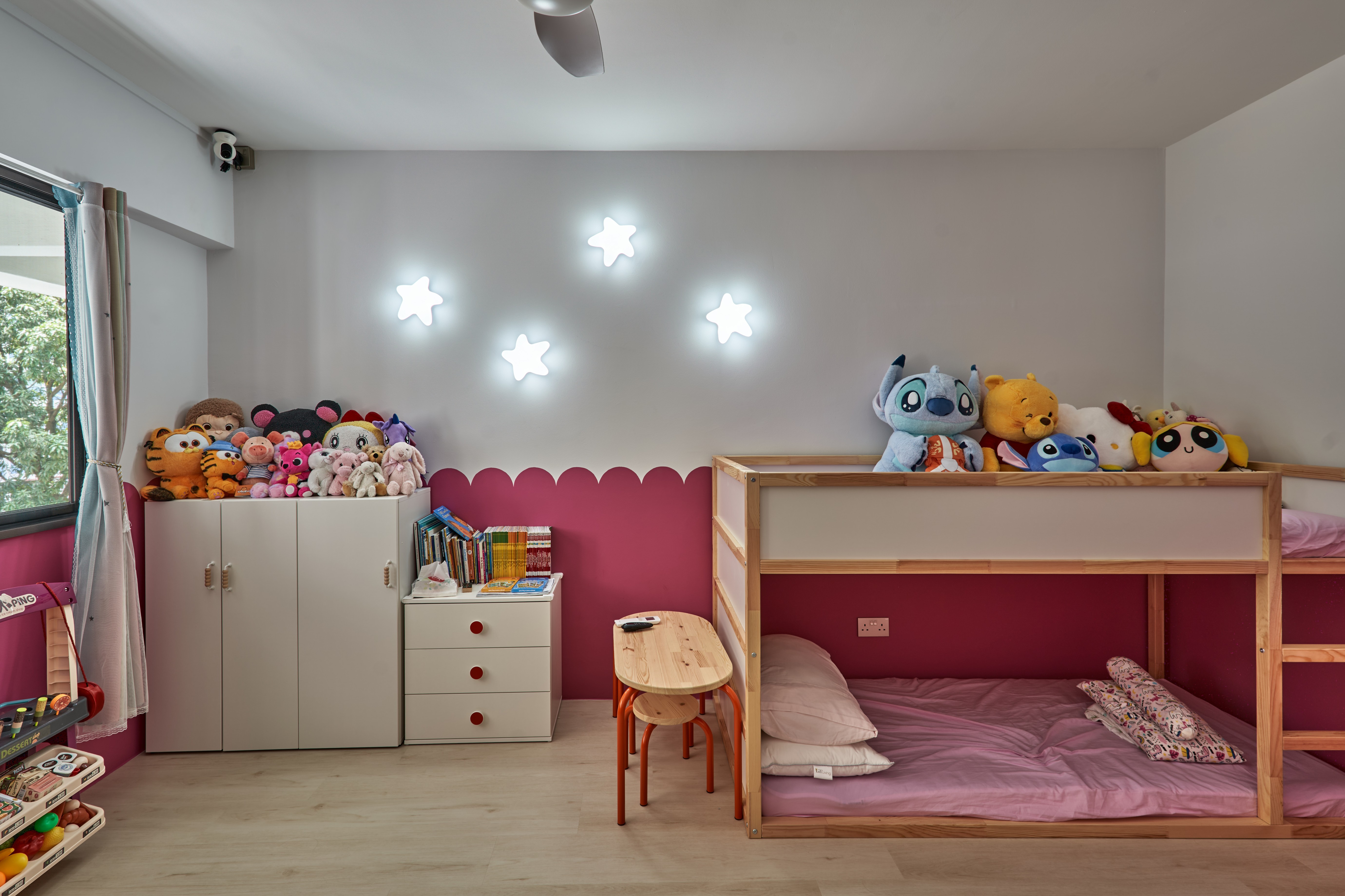 Children's bedroom with a berry-pink accent wall, star-shaped wall lights, white wardrobe, and a wooden bunk bed with stuffed animals and pink bedding.