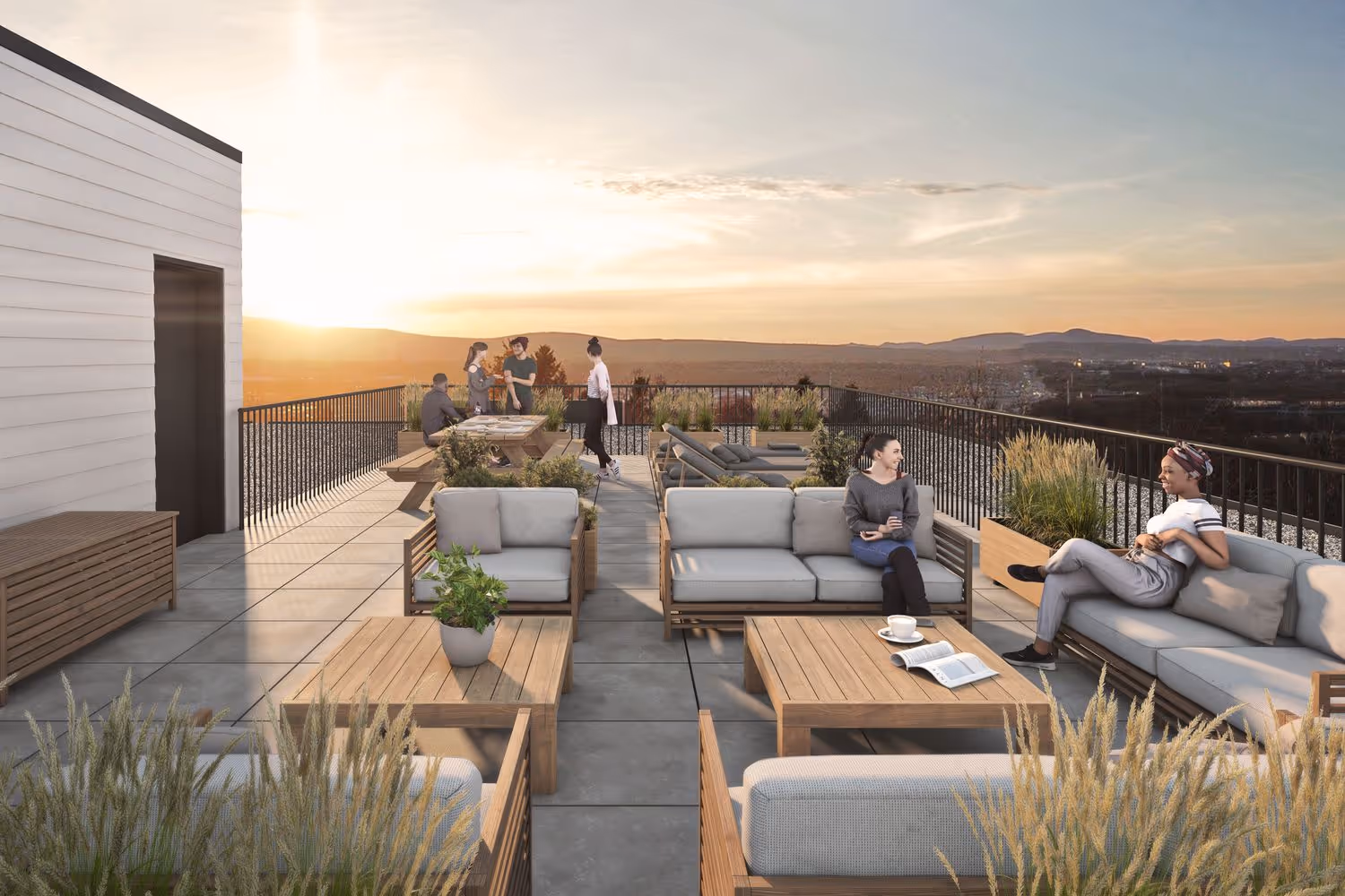 Modern rooftop terrace at sunset with groups of people sitting and chatting on gray cushioned outdoor sofas and wooden tables, surrounded by planters with tall grasses and a panoramic view of distant hills and cityscape.