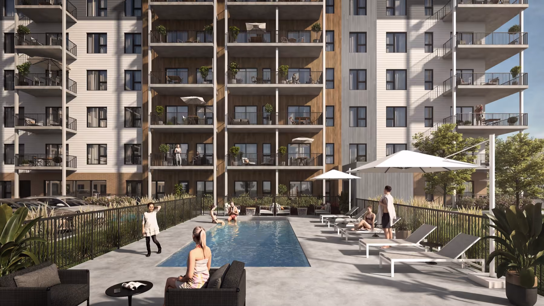Outdoor pool area in front of a multi-story apartment building with balconies, featuring people sitting, standing, and enjoying the poolside seating and lounge chairs shaded by umbrellas.