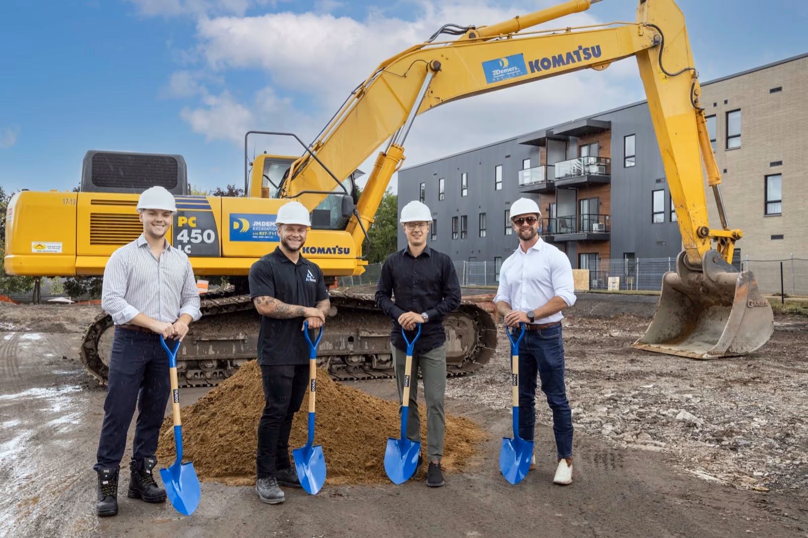 Four construction workers wearing white hard hats standing in front of a yellow Komatsu excavator on a dirt construction site, each holding a blue shovel, with a modern apartment building in the background.