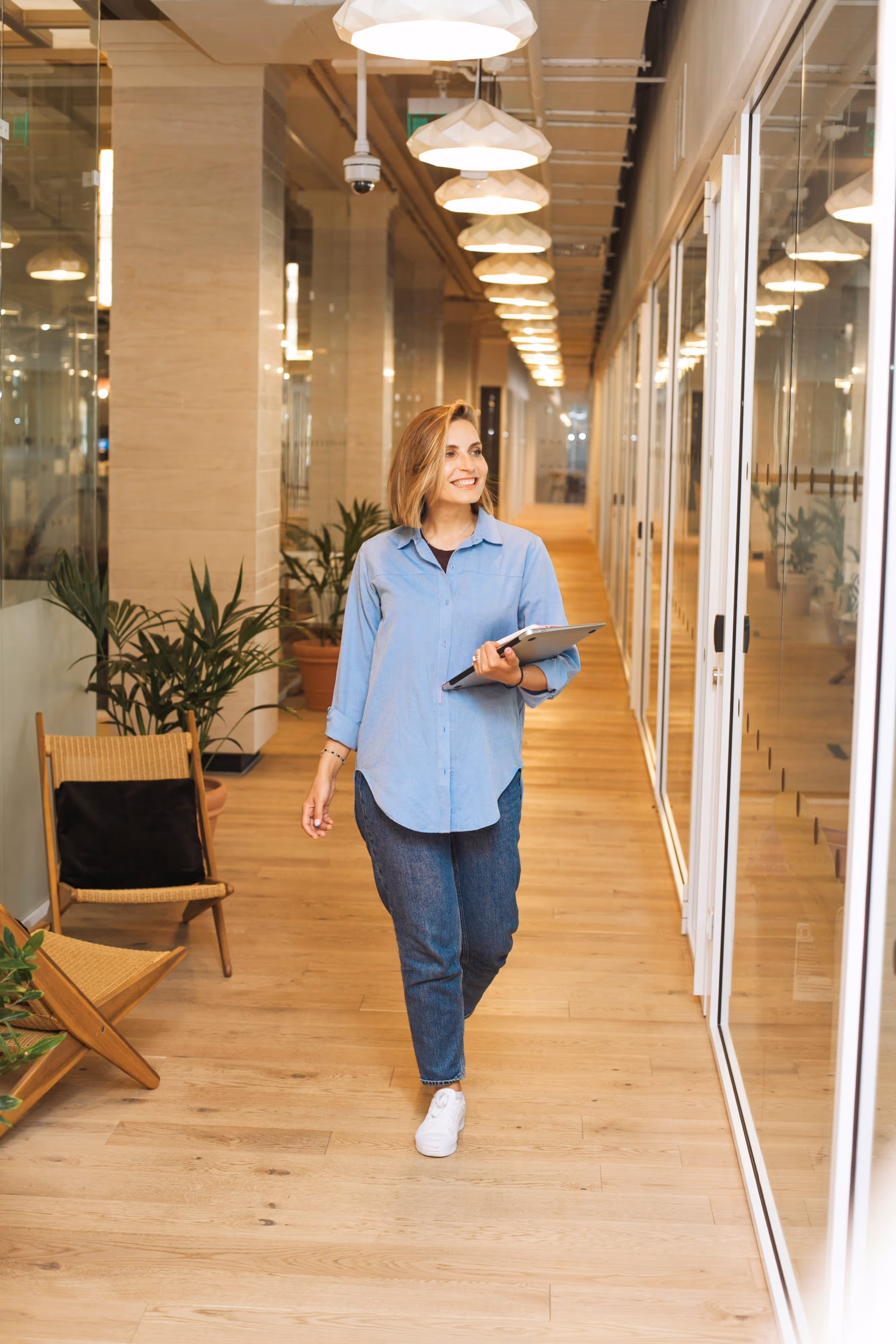 Stock photo of a professional woman smiling and walking down a modern office hallway carrying a tablet and notebook
