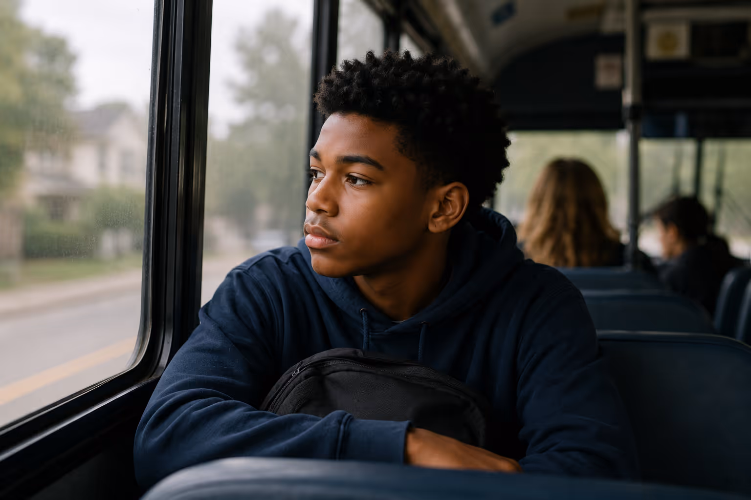 Photo of a teenage boy sitting on a school bus, looking out the window with a backpack on his lap