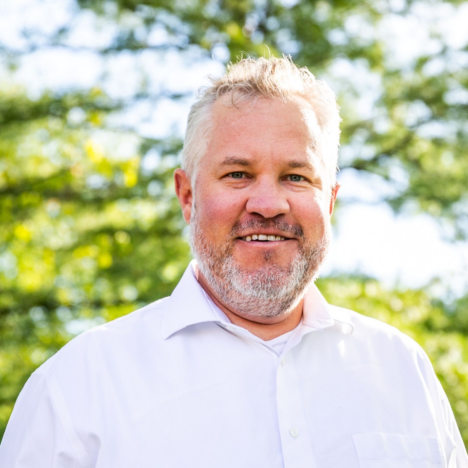 Smiling middle-aged man with gray hair and beard wearing a white shirt outside with green trees.