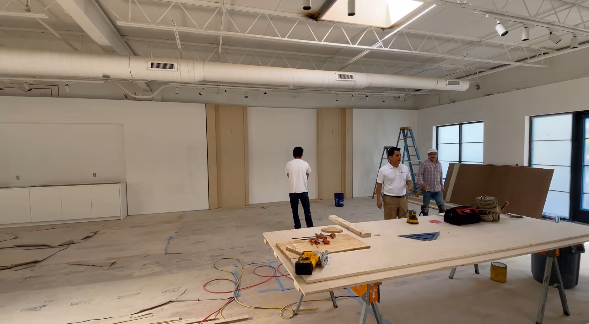 Three men in a large unfinished room with construction tools and materials on a table and floor.