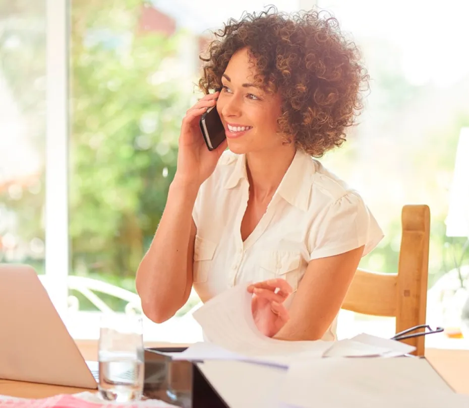 Smiling woman talking on phone while working with papers at a wooden table with laptop and glass of water.
