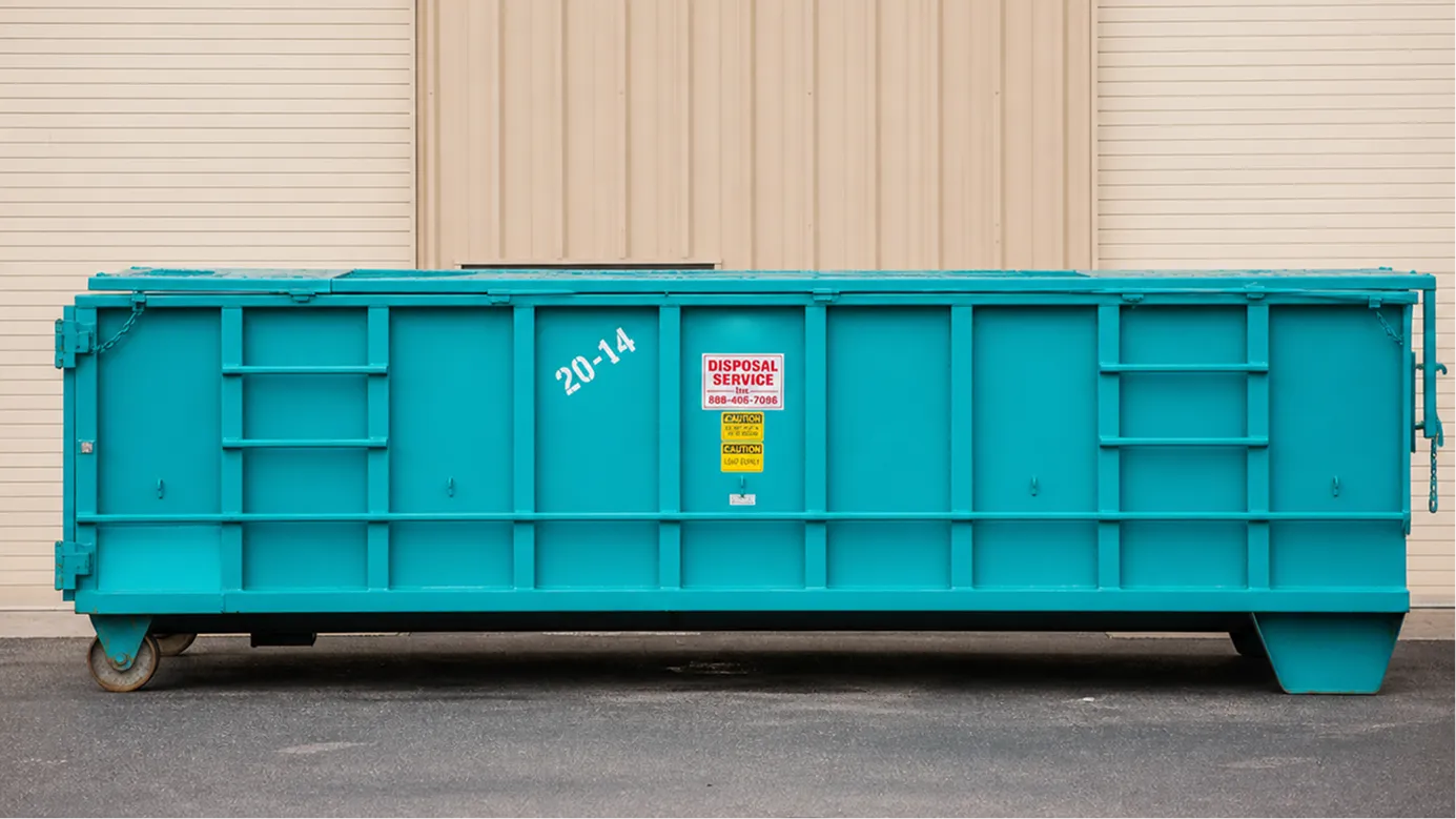 Large turquoise roll-off dumpster against beige building wall on asphalt ground.