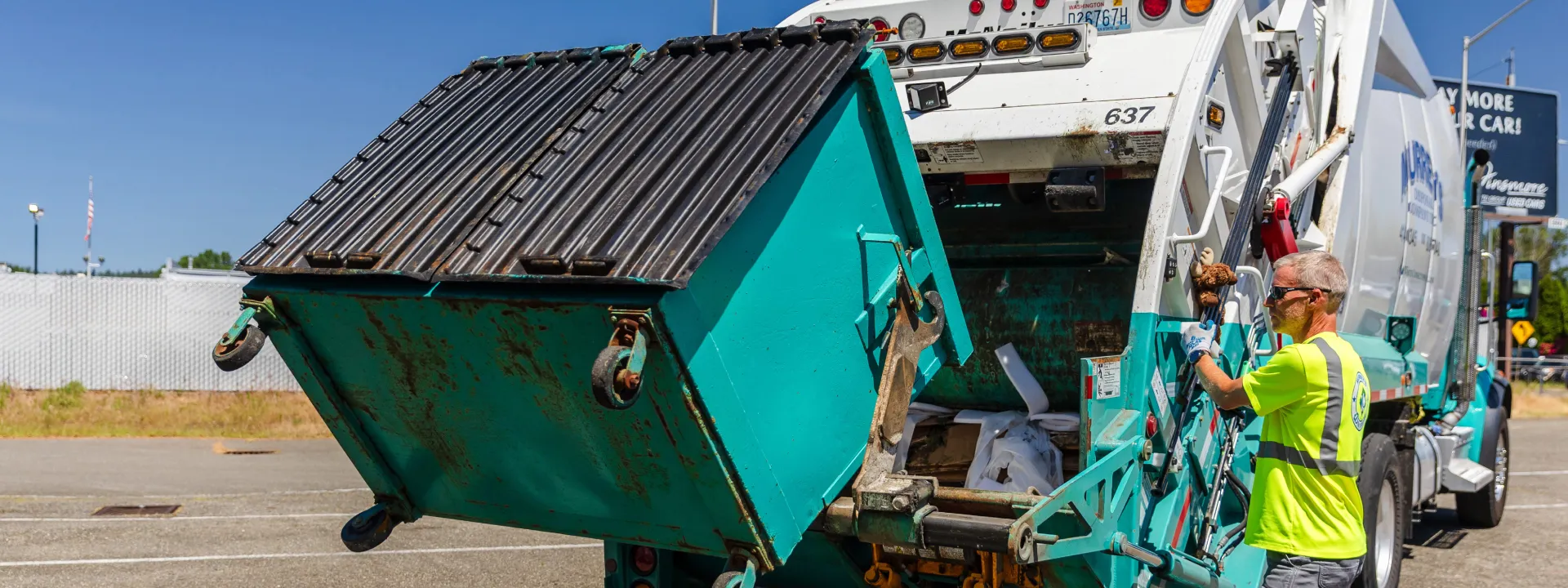 Worker in neon shirt operating truck lifting a large green commercial dumpster outdoors.