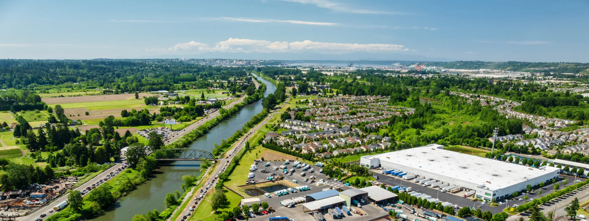Aerial view of a river flowing past a waste service yard, residential area, and green fields under blue sky.