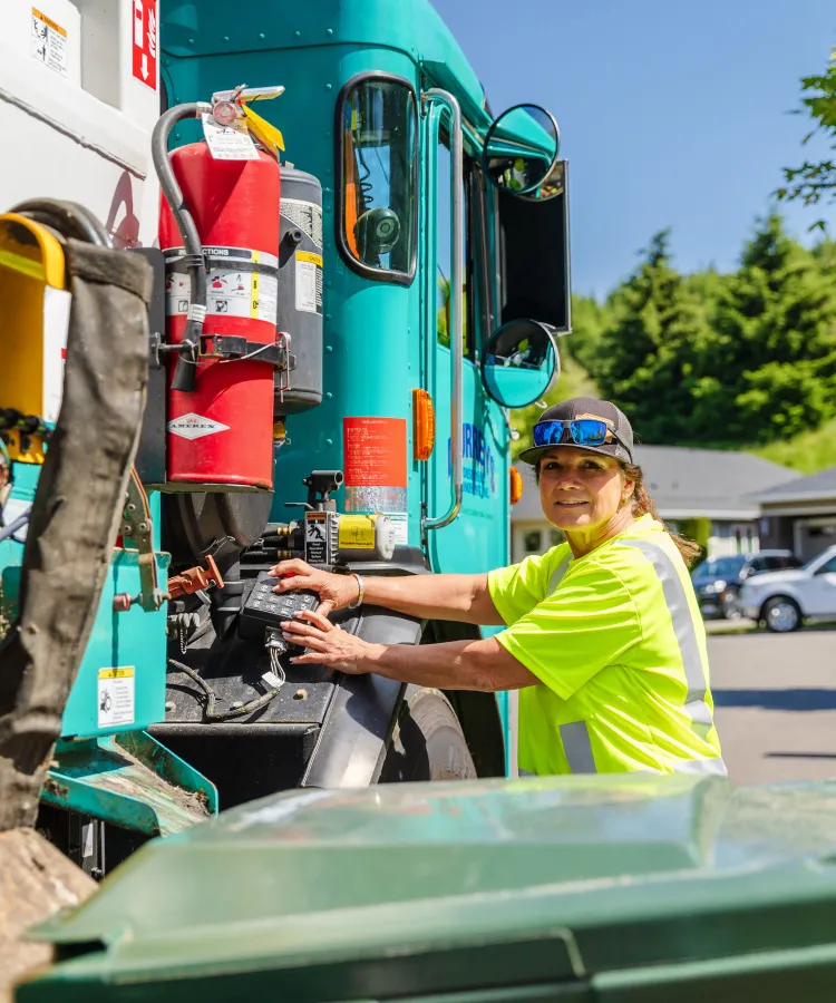 Sanitation worker in a yellow shirt operating controls on a green garbage truck outdoors.