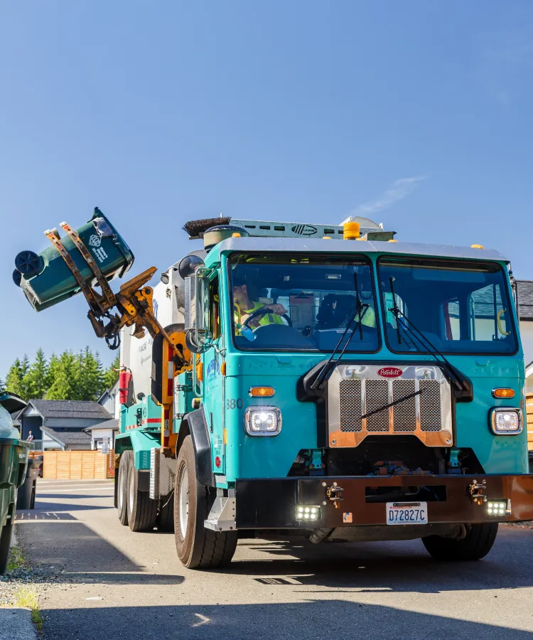 Teal garbage truck lifting a green bin to empty it in a residential driveway on a sunny day.