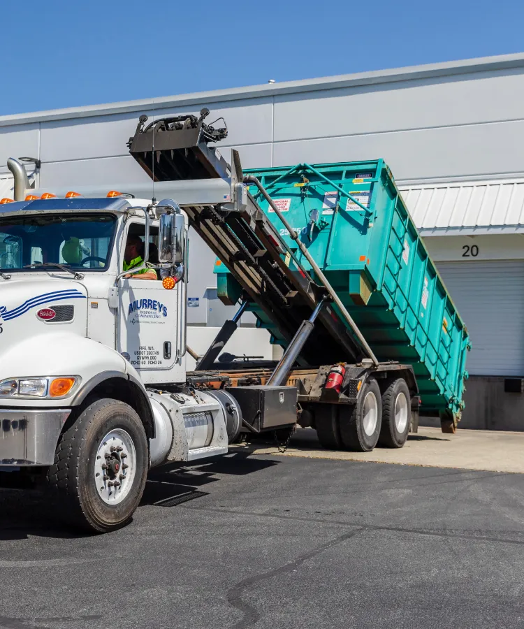 White roll-off truck lifting a green dumpster at a warehouse loading dock under blue sky.
