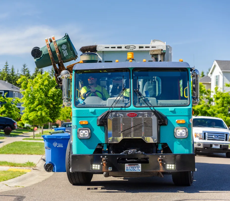 Residential garbage truck lifting a green trash cart to empty into the truck on a sunny neighborhood street.