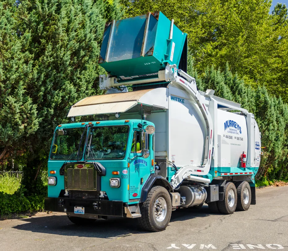 Front-load garbage truck emptying a green dumpster over the cab on a street near trees.