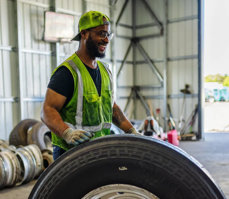 Worker wearing a green safety vest and cap handling a truck tire inside a garage.