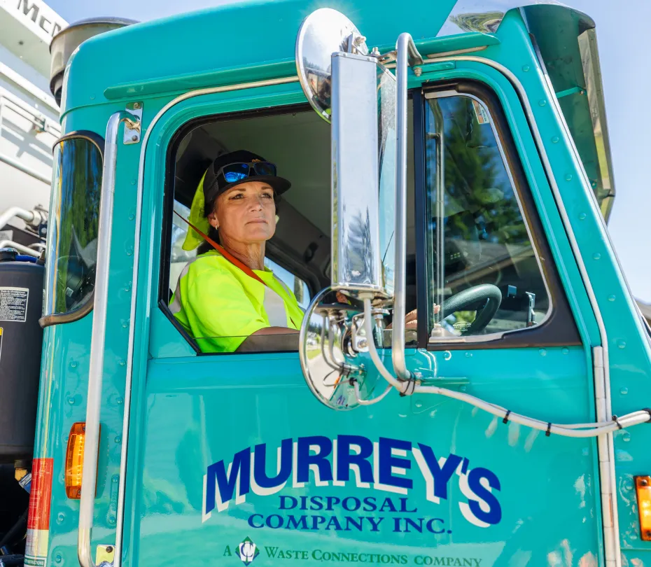 Woman wearing a safety vest and cap sitting in the cab of a teal Murrey's Disposal truck.