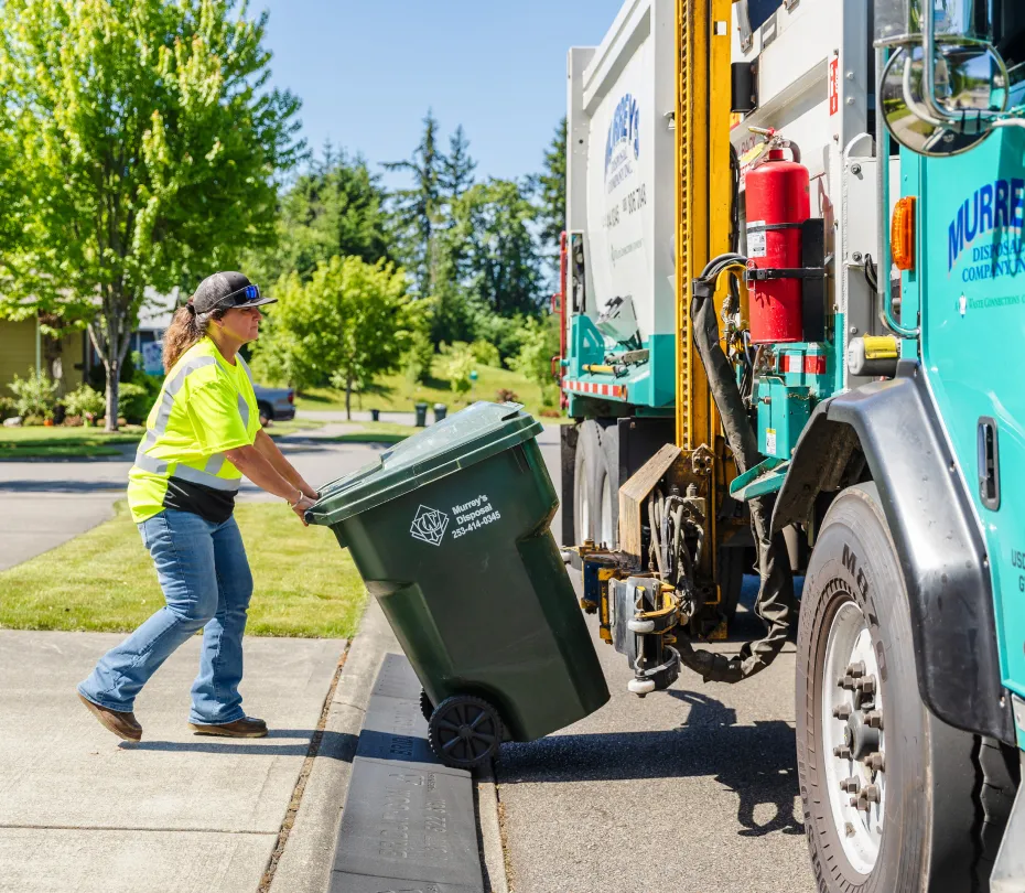 Worker in yellow shirt rolling green residential trash cart toward a collection truck on a sunny street.