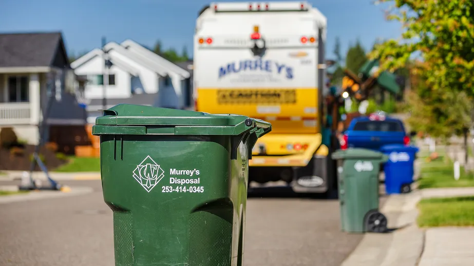 Green curbside trash bin labeled Murrey's Disposal with garbage truck and houses in the background.